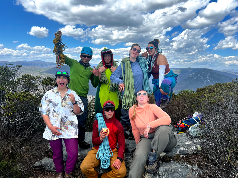 A group of seven people are posing for a photo on a rocky mountaintop. They appear to be climbers, as some are holding ropes and wearing climbing gear. The background features a scenic view of mountains and a partly cloudy sky. The group is dressed in colorful outdoor attire, and some are making playful faces for the camera.